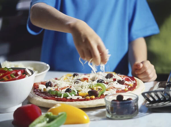 Cheese being sprinkled over pizza with vegetable toppings, surrounded by olives and sliced peppers in bowls, spatula