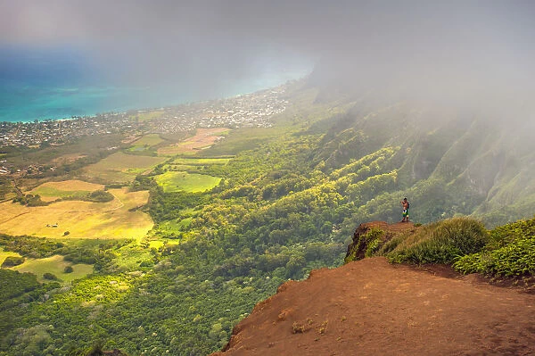A Man Standing On The Edge Of A Cliff On The Kuliouou Ridge Trial Enjoys The View Of Oahus Windward Side And The Town Of Waimanalo As The Clouds Roll In On A Summers Day; Waimanalo, Oahu, Hawaii, United States Of America