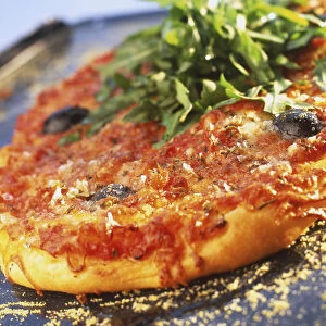 Round pizza with thick chewy crust, topped with tomato sauce, cheese, herbs, two whole olives and garnished with ruccola leaves in middle, tilted view