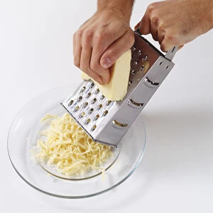 Using metal grater to manually grate cheddar cheese onto a clear glass plate, high angle view
