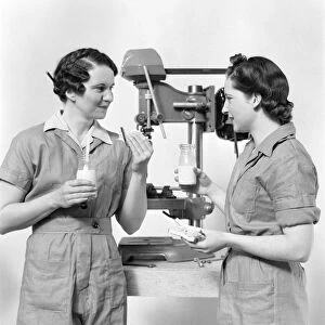Two women workers standing near drill press, eating sandwich and drinking milk