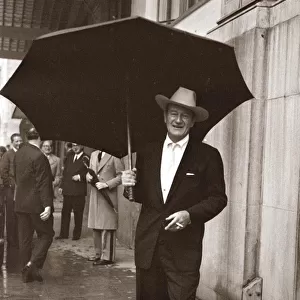 John Wayne standing under a big black umbrella in the rain in London October 1960